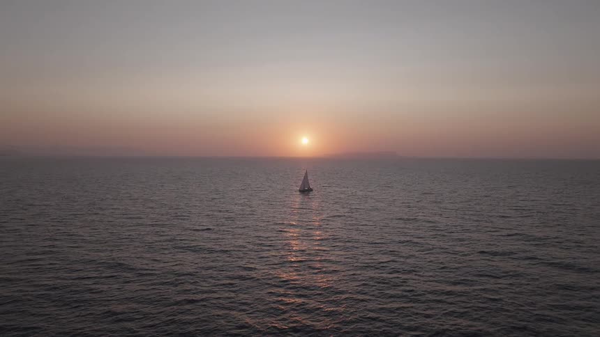 A lone sailboat with a white sail is sailing on a vast dark ocean as the sun sets casting a long shimmering reflection of light on the water