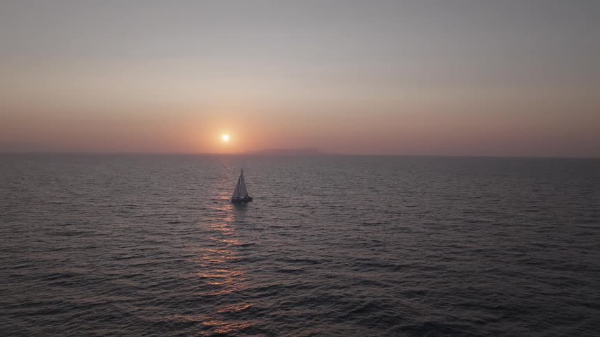 A wide shot of a sailboat sailing peacefully on the open ocean silhouetted against a brilliant sunset