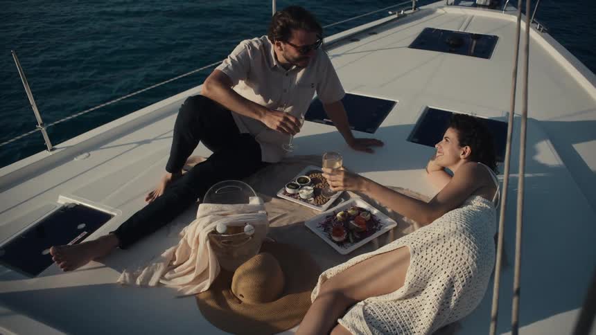 A man and a woman are relaxing on the deck of a boat with food and drinks laid out on a tray between them the sun is shining brightly creating reflections on the deep blue water surrounding the boat