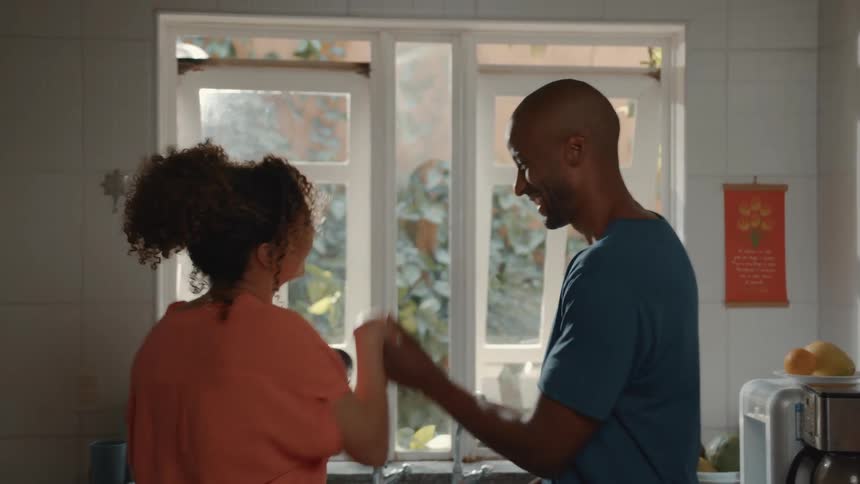 A smiling couple is playfully dancing together in a brightly lit kitchen with the woman wearing an orange shirt and a man in a blue tshirt with his arms wrapped around her