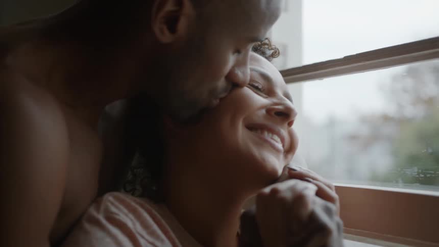 A couple cuddles by the window indoors smiling and enjoying each others company