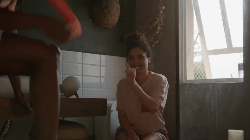 A candid moment of a couples intimate morning routine where a woman sits on a toilet and brushes her teeth while talking to her partner