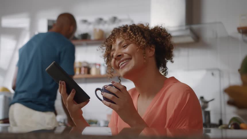 A woman is sitting at a kitchen counter happily looking at a tablet while sipping from a mug with a man visible in the background