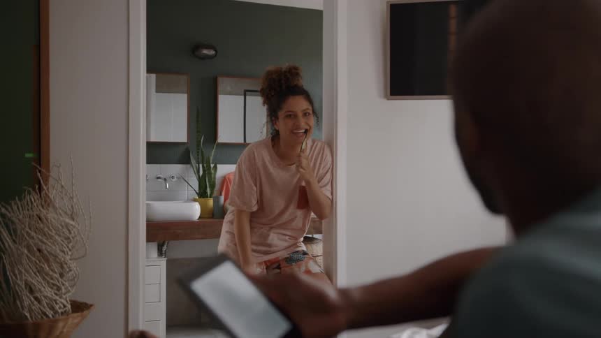 A woman brushes her teeth while talking to a man inside the bathroom