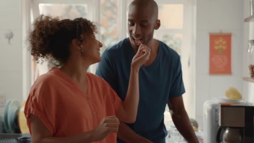 A woman is holding up a small piece of food and offering it to a man they are in a kitchen smiling and interacting playfully