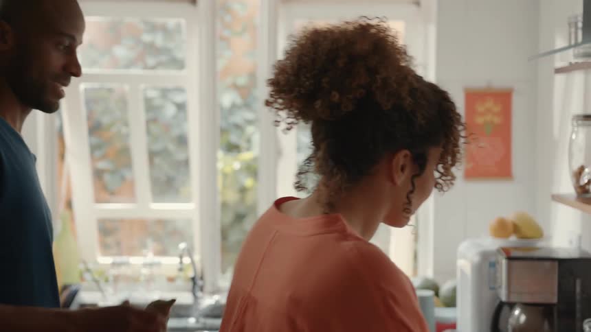 A happy couple stands together in a kitchen with a woman in an orange shirt laughing at her partner a man in a blue tshirt who helping her prepare food