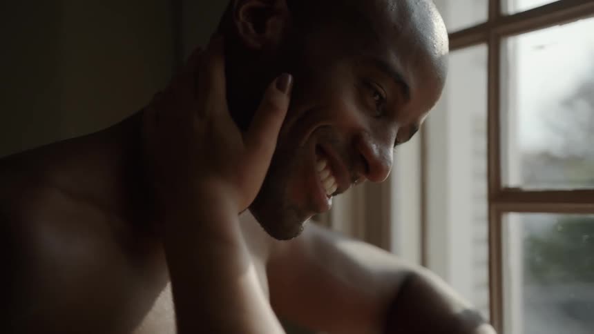 A happy smiling shirtless man is sitting in front of a window and leaning forward as a womans hand caresses the side of his neck and face