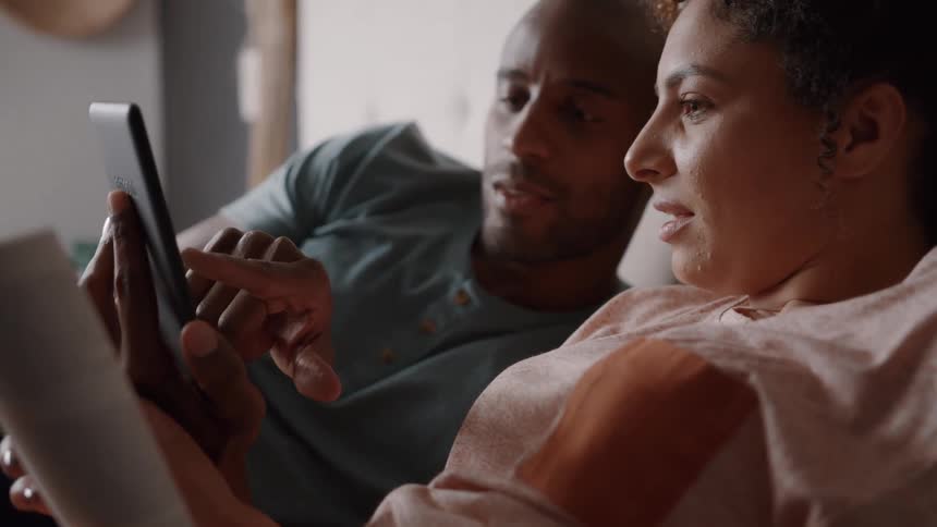 A person is holding a book and pointing at a tablet the person is sitting on a couch next to a man who is watching the tablet
