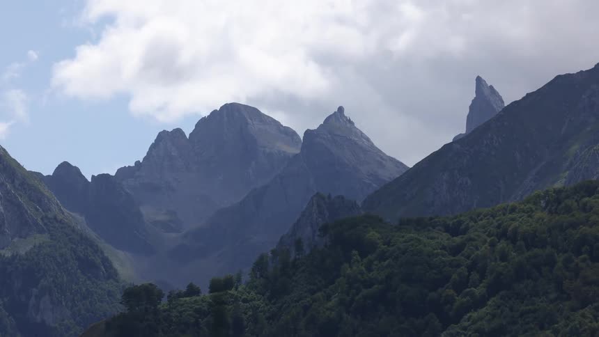 A rugged mountain range covered in green trees as a sunny blue sky with white clouds gives way to an overcast and gloomy day
