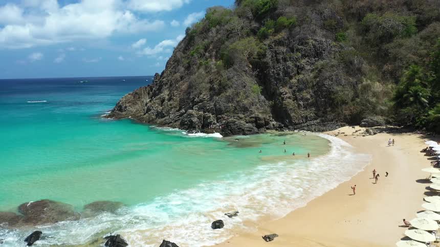 An aerial view reveals a stunning tropical cove with turquoise water where people swim and relax on the sandy beach next to a large rocky hill