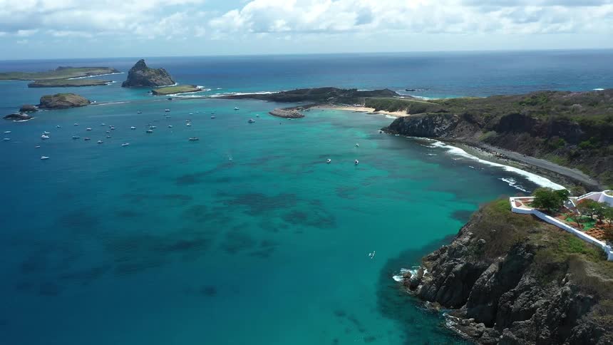 A large bay with turquoise water is shown from above with many sailboats scattered across the water a white building on a rocky cliff and a line of palm trees visible on the shore