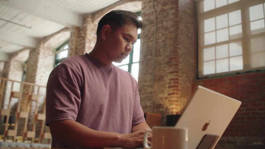 A professional man in a pink tshirt is sitting at a wooden table in a brightly lit room with a high ceiling and exposed brick typing on a laptop with a mug next to him