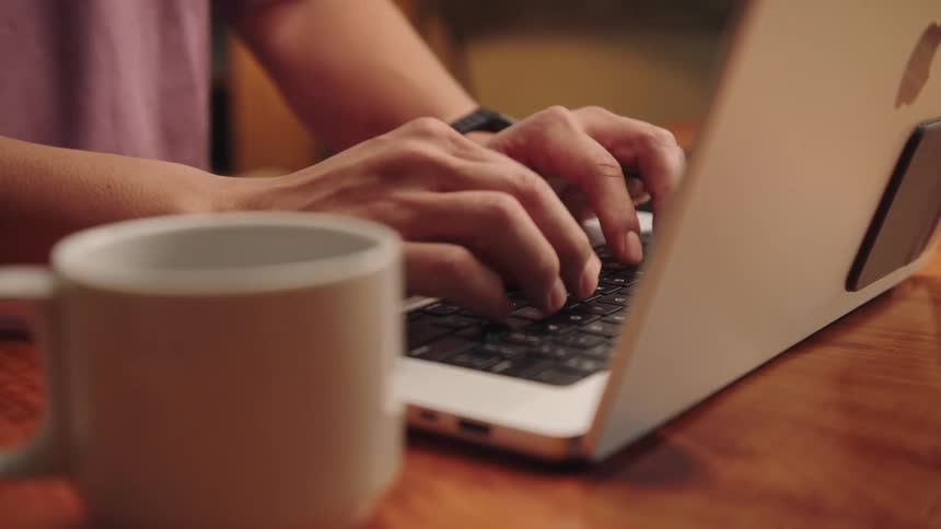 A mans hands are shown typing on a laptop keyboard a mug next to the computer suggesting a remote work setting
