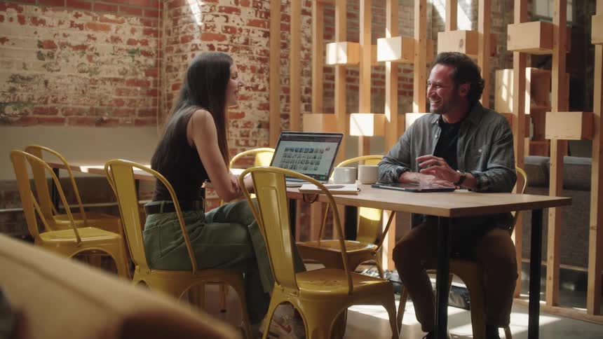 A woman with long dark hair and a man in a gray shirt and jeans are having a discussion while sitting across from each other at a table with a laptop in a brightly lit room with exposed brick walls