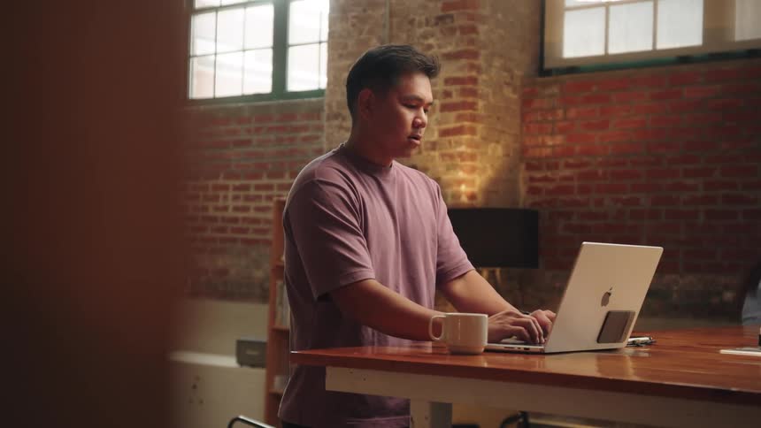 A person stands at a high wooden table in a room with a brick wall and large windows they are typing on a laptop and holding a white mug