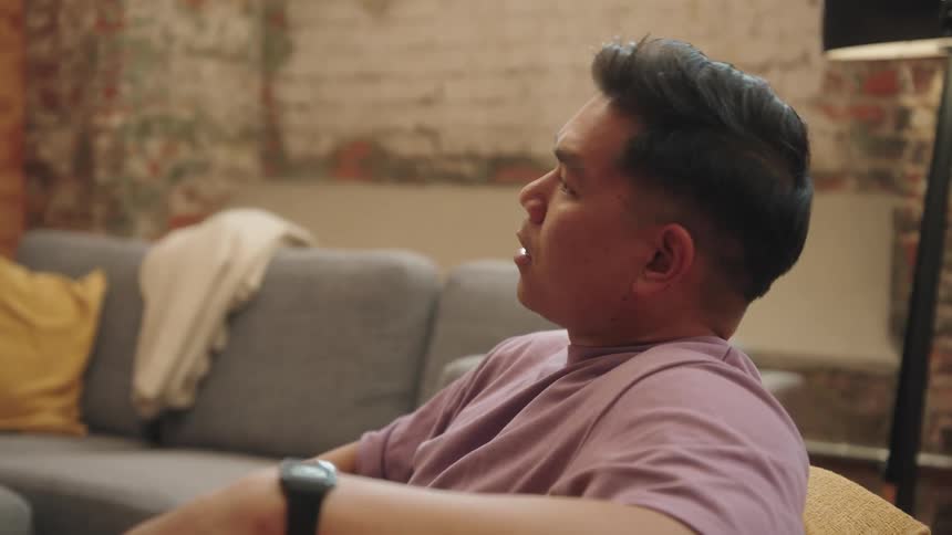 A man with dark hair is sitting on a couch in a modern office space he is looking offcamera and has his arm resting on the back of the couch