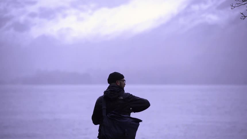 A man is seen from behind standing in front of a large body of water under a cloudy sky he is wearing a dark jacket with a hood and a beanie