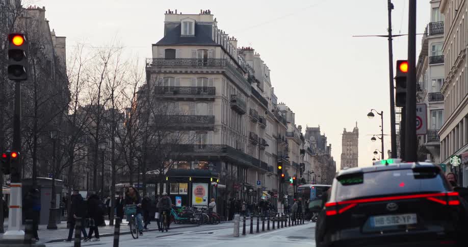 A busy city street during the day capturing the flow of pedestrians and vehicle traffic with ornate multistory buildings lining both sides of the street