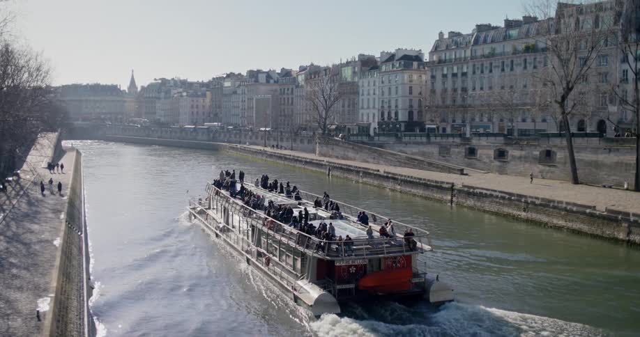 A riverboat filled with tourists travels along a river lined with historic buildings the boat with a clear upper deck leaves a wake behind it as it moves through the water on a sunny day