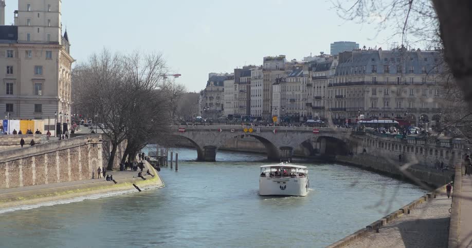 A boat travels along a canal beneath a bridge in the city with cars passing by at midday