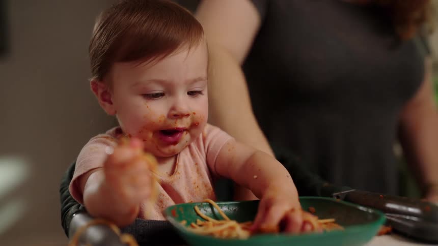 A happy baby is sitting in a high chair eating messy spaghetti with their hands as their mother who is wearing a gray tshirt stands behind them