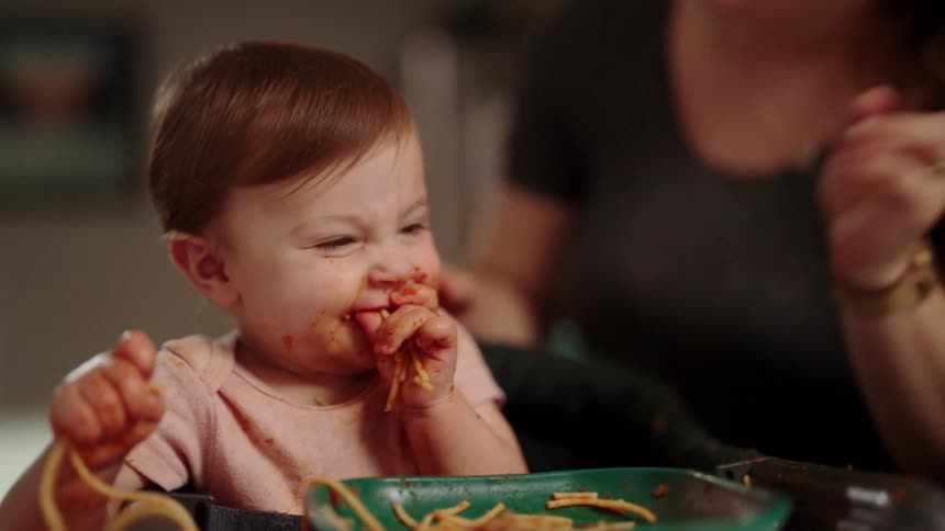A toddler eats a spaghetti meal with his bare hands as the mother comes from behind to kiss him on the cheek both laughing