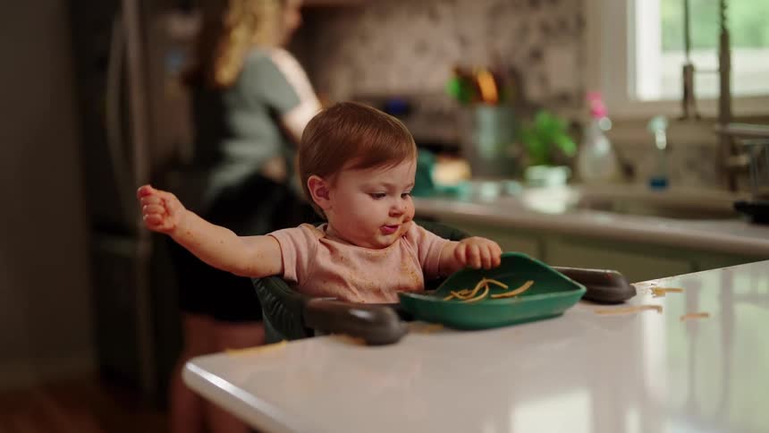 A child is eating from a plate on the kitchen countertop while the mother cooks another meal behind him at midday