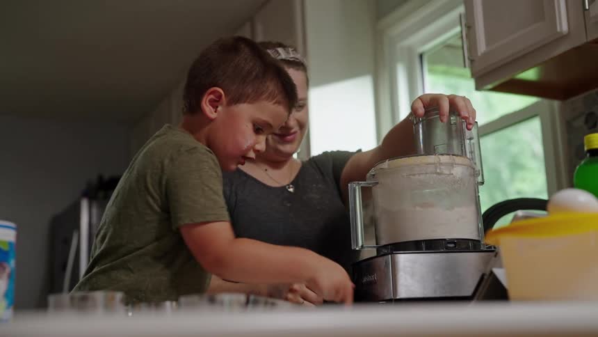 A boy and an adult woman are standing at a kitchen counter using a food processor the woman is holding the lid of the food processor while the boy reaches to help they appear to be making a dough or batter together