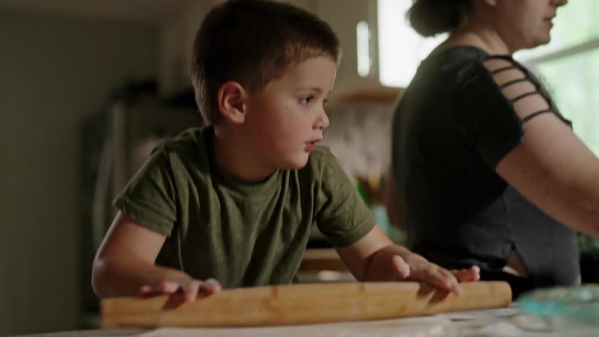 A young boy rolls dough with a wooden rolling pin while a woman works beside him in the kitchen