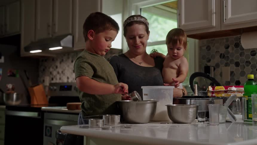 A mother holding a baby is helping her young son scoop white flour into a large metal mixing bowl on a kitchen countertop