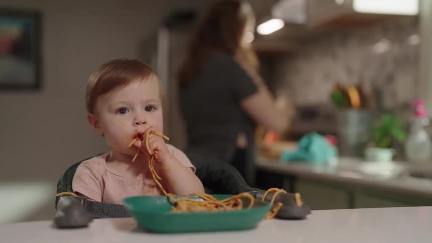 A baby in a high chair happily eating spaghetti with their hands in a kitchen with an outoffocus adult in the background