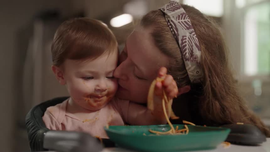 Mother giving a kiss to her little daughter who is eating messy pasta