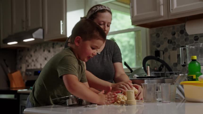 A happy mother and her young son are in a kitchen with the mother helping the boy press a mound of dough on a countertop