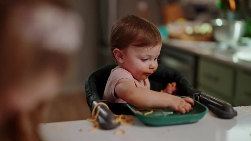 A baby in a high chair reaches for spaghetti on a plate with sauce on their face