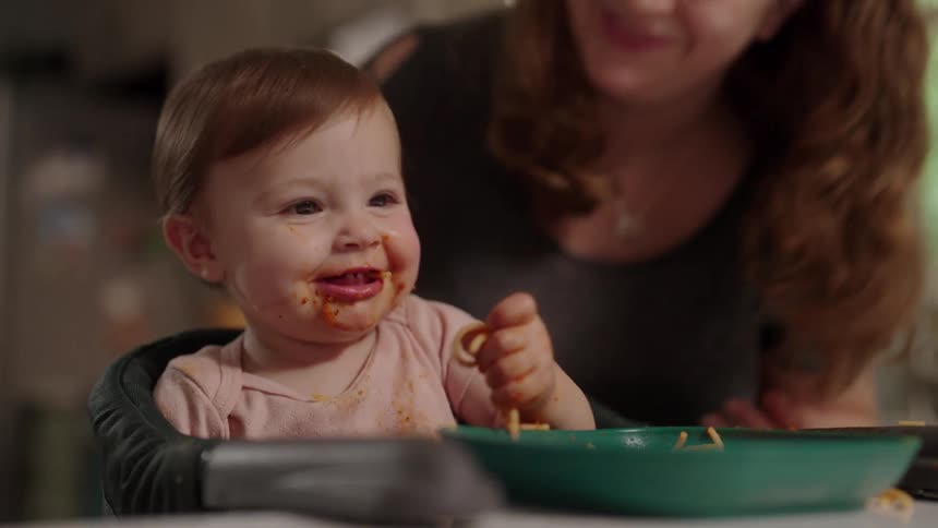 Mother kisses her daughter while the child sits in a highchair in their kitchen and eats messily
