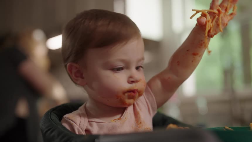 A baby eats spaghetti with sauce covered hands leaning forward into the meal