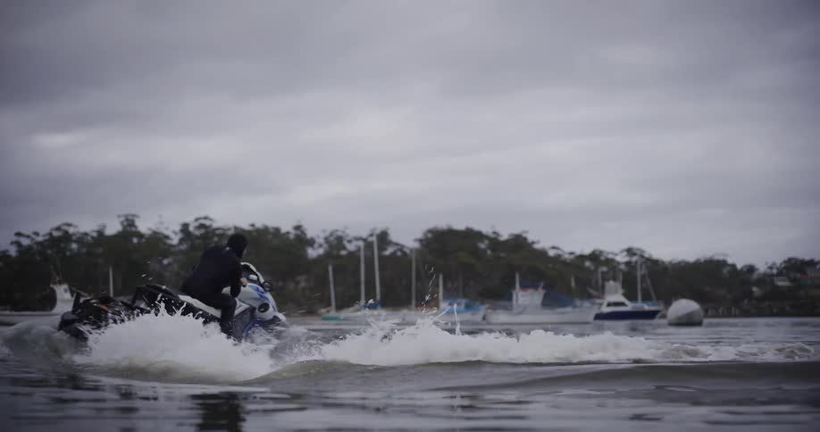 A person in a wetsuit starts their jet ski next to a dock and speeds off across a harbor on an overcast day