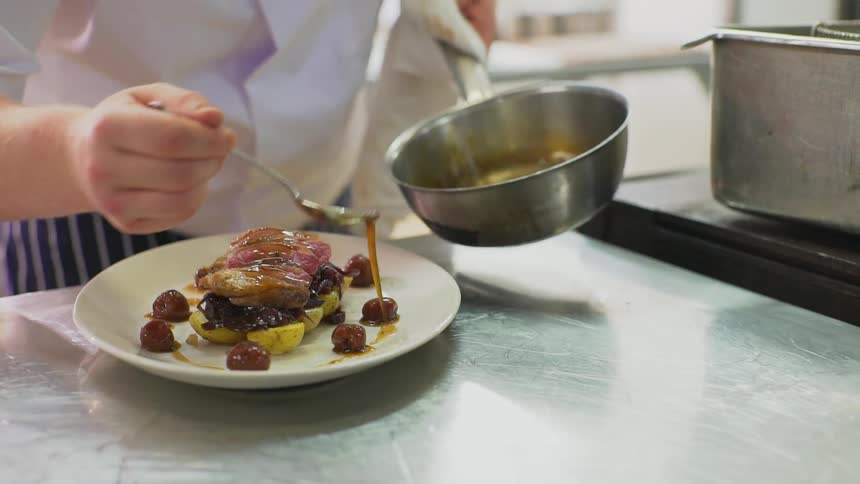 A person pulling liquid out of a pan with a spoon and pouring it over some food on a plate