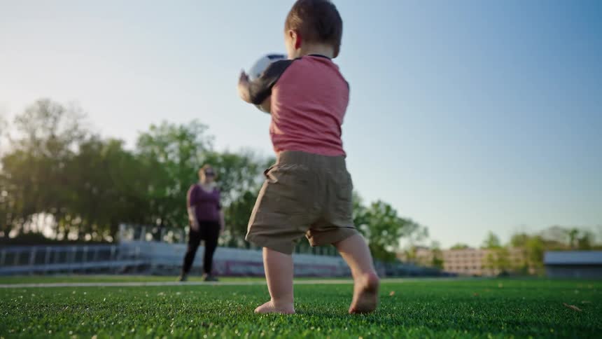 A baby wearing a red and black shirt and tan shorts is standing on a soccer field attempting to pick up a soccer ball as a parent stands in the background