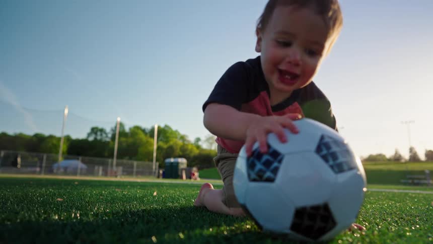A toddler kicks a soccer ball on a sunny field while an adult watches from behind