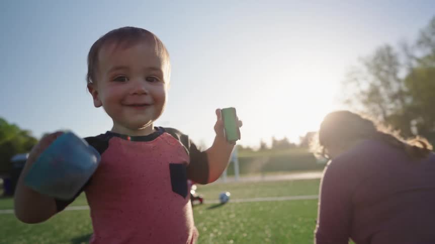 A child playing with a toy on a football field walks by his mother sitting on the ground at midday
