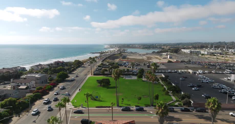 A slow aerial shot pans across a sunny coastal town showcasing a road running parallel to the beach a green park and houses