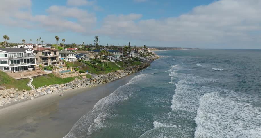An aerial view glides along a sunny coastline revealing upscale homes atop a cliff overlooking a sandy beach with breaking ocean waves dotted with surfers under a blue sky