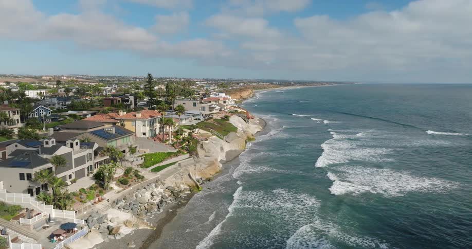 A large body of blue ocean water with white waves crashes against a sandy beach bordered by a steep rocky cliff with many houses built along the top