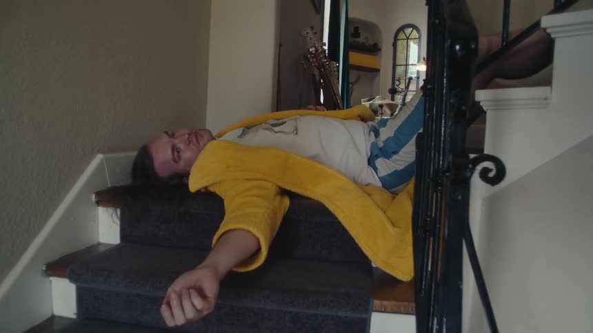 A man wearing a yellow jacket lies on the staircase appearing to have fallen and landed on his back indoors at midday
