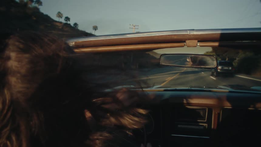 The video shows a firstperson view from the drivers seat of a car as it travels along a treelined road the drivers hand is visible on the steering wheel and the rearview mirror hangs from the windshield