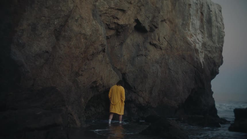 A person in a bright yellow robe standing on a dark wet shoreline with their back to the camera facing a massive craggy rock formation as gentle waves lap at their feet