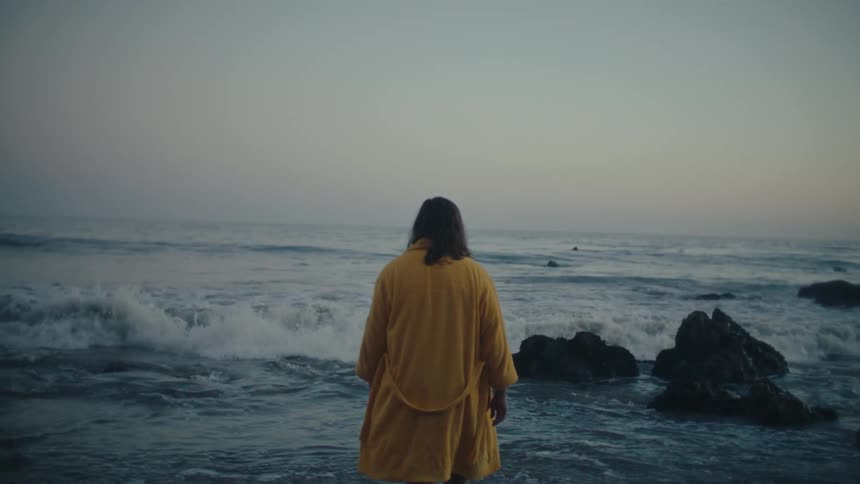 A longhaired person in a yellow robe stands facing the sea with their back to the camera the person is wading in the shallow water with waves breaking on the shore around their feet