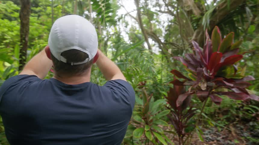 A man wearing a white baseball cap a dark blue tshirt and black shorts is standing on a paved path in a lush botanical garden using his smartphone to take a picture of a large red and green plant