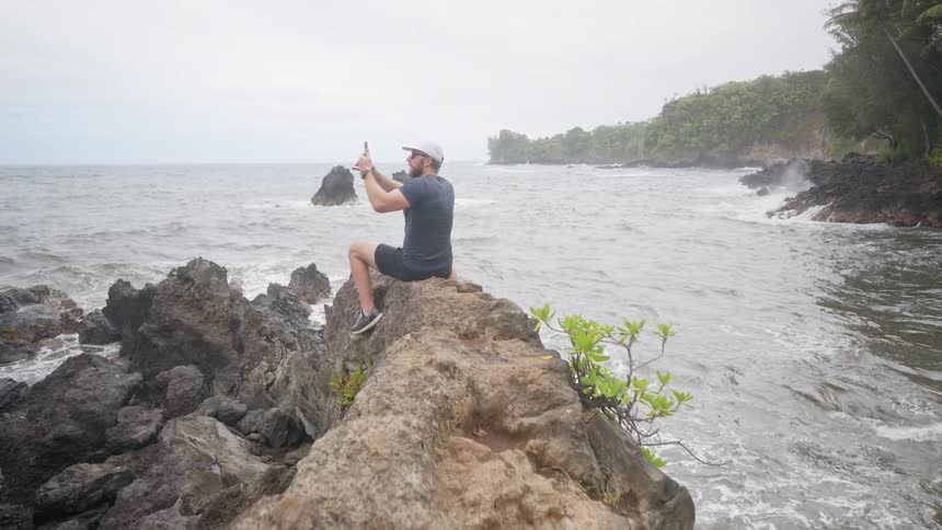 A man in a white cap sits on coastal rocks photographing the ocean on an overcast day